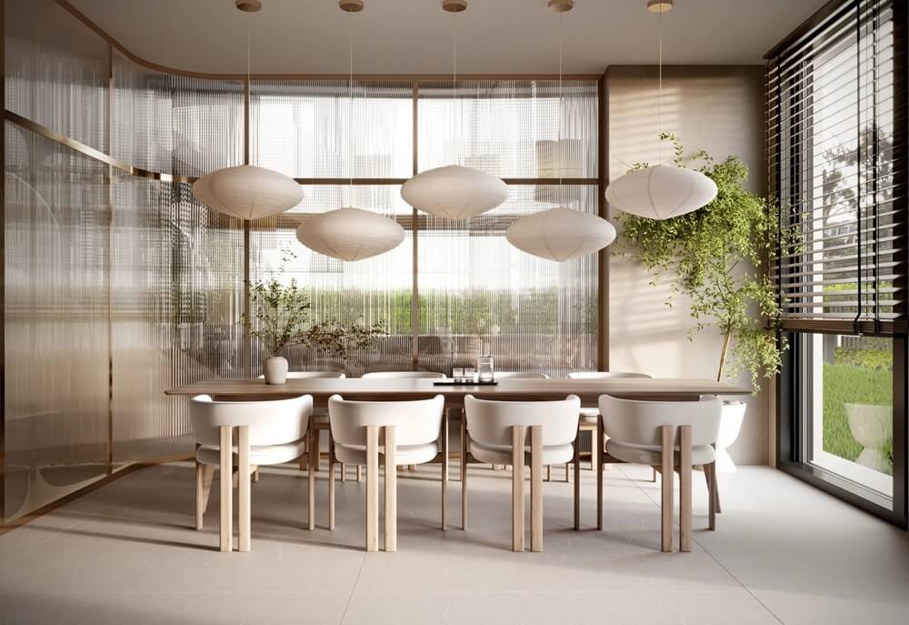 Ця сучасна їдальня випромінює елегантність та спокій. Центральним елементом є довгий прямокутний стіл зі світлого дерева, оточений вісьмома білими стільцями з мінімалістичним дизайном. Над столом висять сім великих овальних ламп, створюючи вражаючу світлову композицію та підкреслюючи сучасний характер простору. Фоном є ніжні напівпрозорі штори та великі вікна з горизонтальними жалюзі, що відкривають вид на зелений сад. Дві високі рослини в горщиках додають інтер'єру природного та свіжого відчуття.