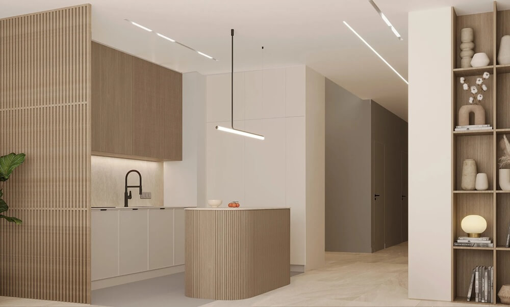 照片展示了一间与走廊相连的现代简约厨房。厨房以浅色调为主,白色柜门与原木材质相得益彰,营造出温馨优雅的氛围。厨房的视觉焦点是一个木质小岛台,上方悬挂着一盏纤细的线性吊灯。左侧的竖条木条增添了纹理感和舒适感。右侧的嵌入式书架摆放着书籍、花瓶和台灯等装饰品,更添一份居家气息。通往其他房间的走廊则由一盏低调的吸顶灯照亮。